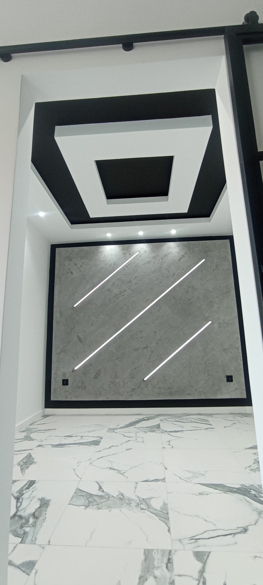 Nowoczesne wykończenie wnętrza: szara ściana z podświetleniem LED, marmurowe płytki podłogowe i designerski sufit podwieszany z czarno-białymi elementami.