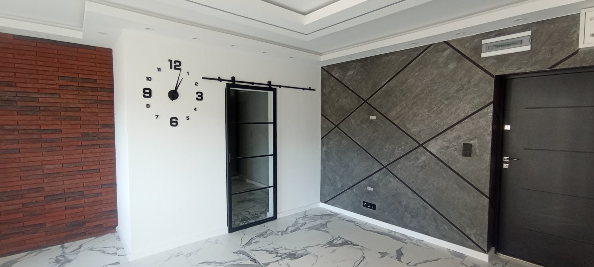Nowoczesne wykończenie wnętrza w Łasku: ceglana ściana, dekoracyjna szara ściana z geometrycznym wzorem, przesuwne drzwi i duży zegar na ścianie.