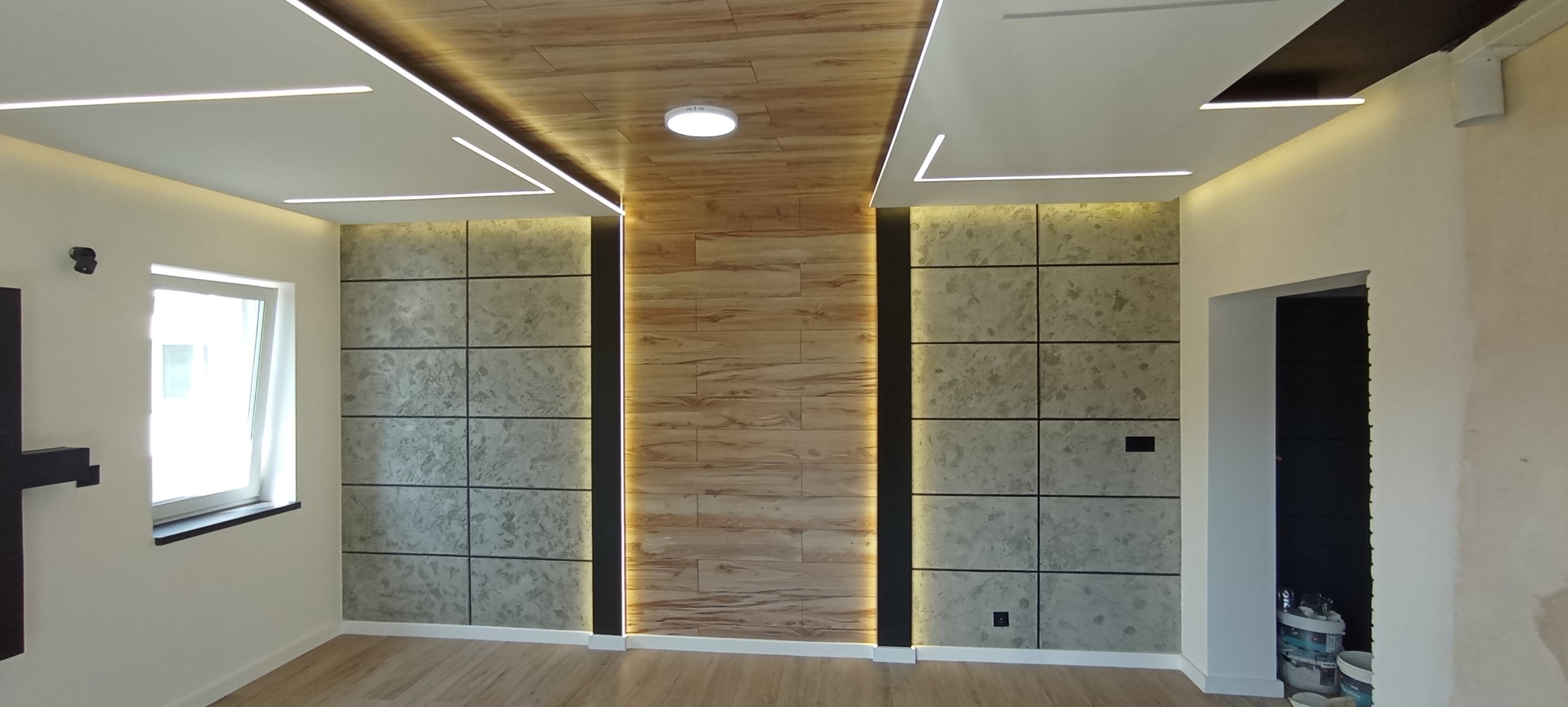 Nowoczesne wykończenie wnętrza: panele dekoracyjne na ścianie, drewnopodobny sufit z oświetleniem LED, jasna podłoga. Widoczne detale wykończeniowe i elementy instalacji.
