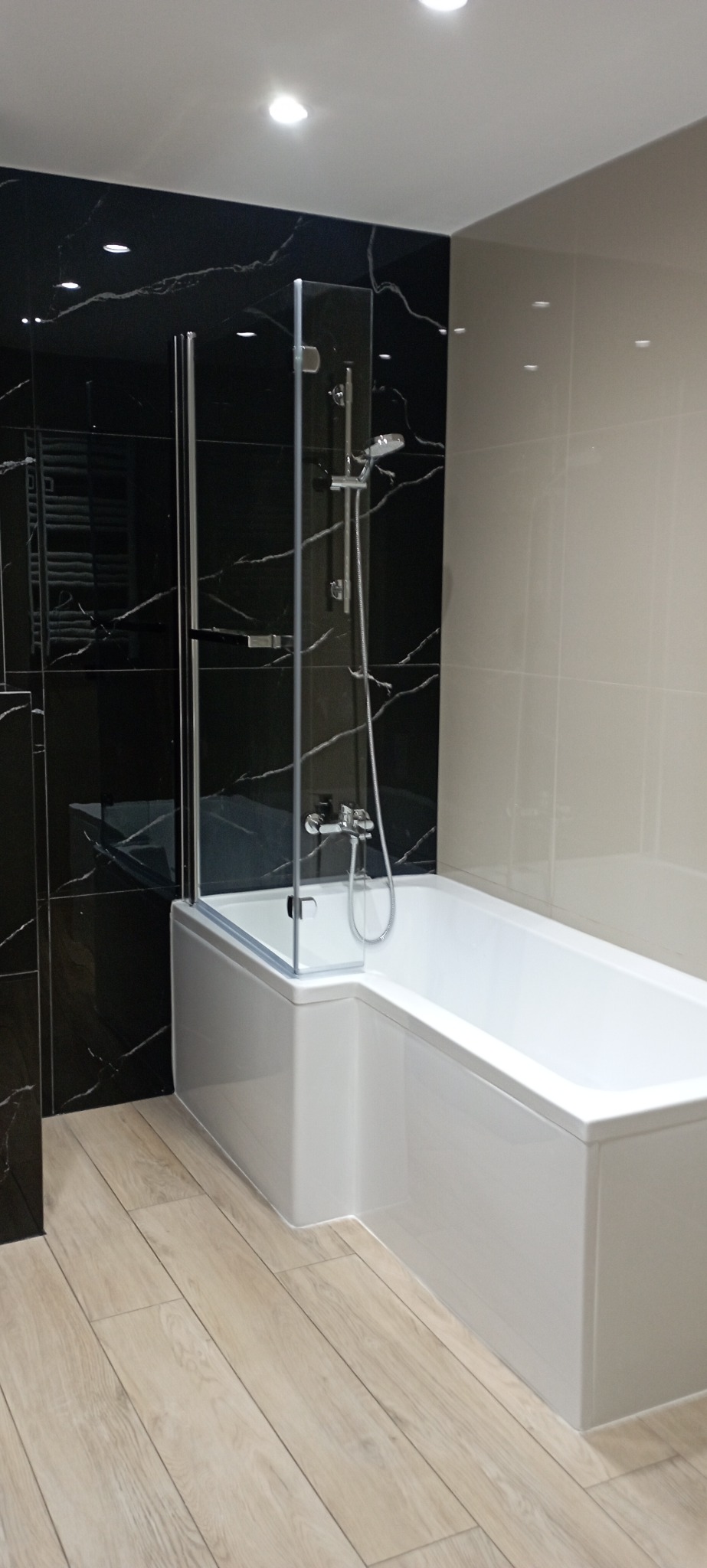 Nowoczesna łazienka z wanną i szklanym parawanem, czarne płytki imitujące marmur na ścianie, jasna podłoga z paneli. Minimalistyczny design.
