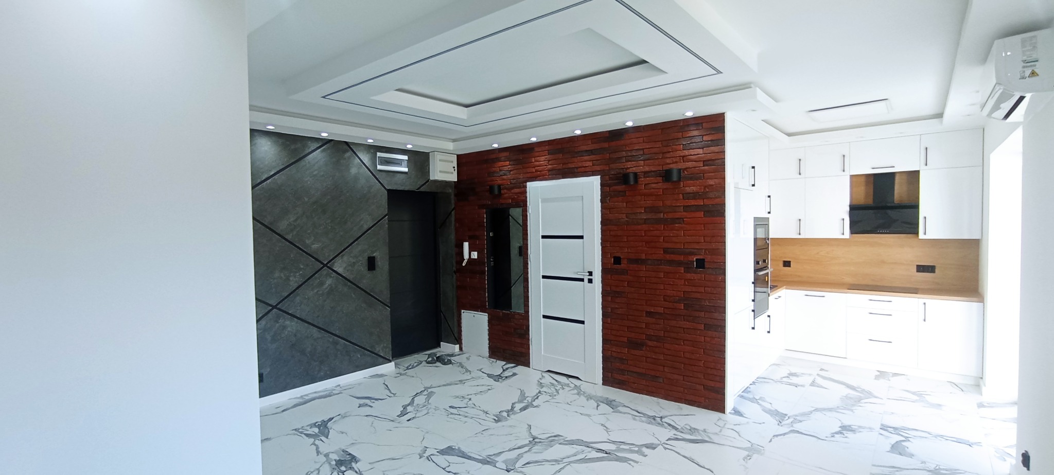 Nowoczesne wnętrze: grafitowa ściana z geometrycznym wzorem, ceglana ściana z białymi drzwiami i fragment białej kuchni z drewnianym blatem. Marmurowe płytki na podłodze.