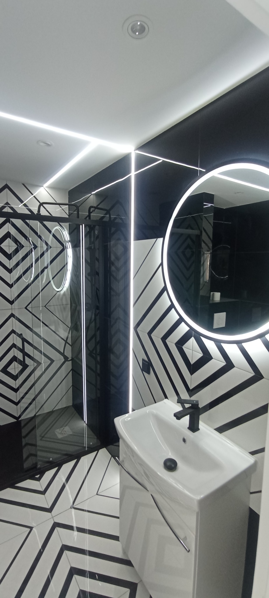 Nowoczesna łazienka w czarno-białej kolorystyce z geometrycznym wzorem na ścianach i podłodze. Umywalka z czarną armaturą, okrągłe lustro z podświetleniem LED i kabina prysznicowa.