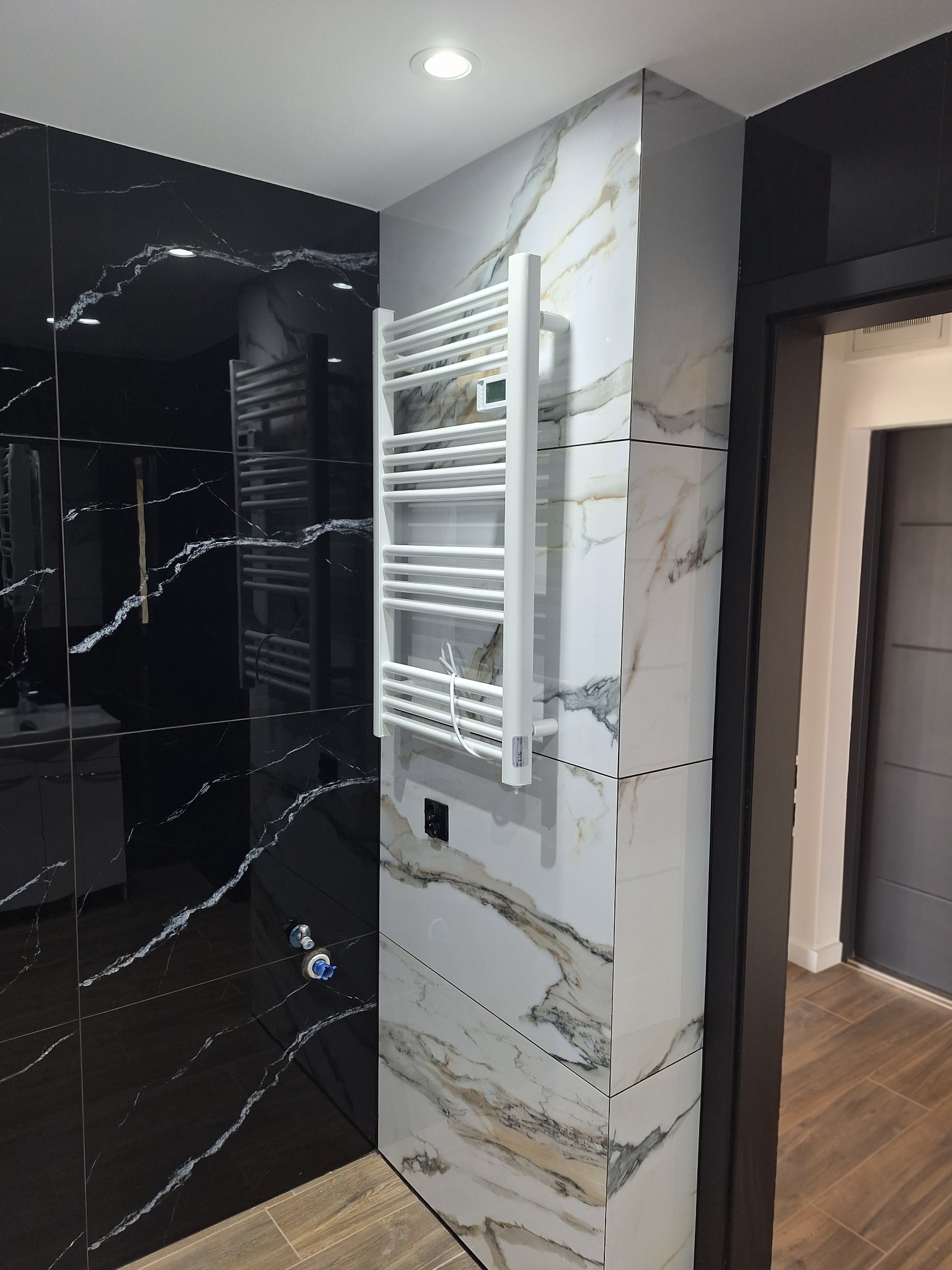 Nowoczesna łazienka z czarnymi i białymi marmurkowymi płytkami, grzejnikiem drabinkowym i drewnopodobną podłogą. Widoczny fragment drzwi.