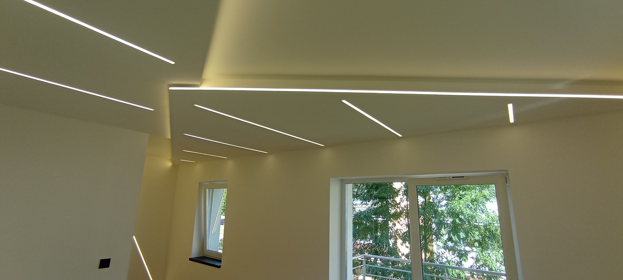 Nowoczesne oświetlenie sufitowe z liniowymi lampami LED w jasnym wnętrzu. Widoczne okna z widokiem na zieleń. Minimalistyczny design, świeżo po remoncie.