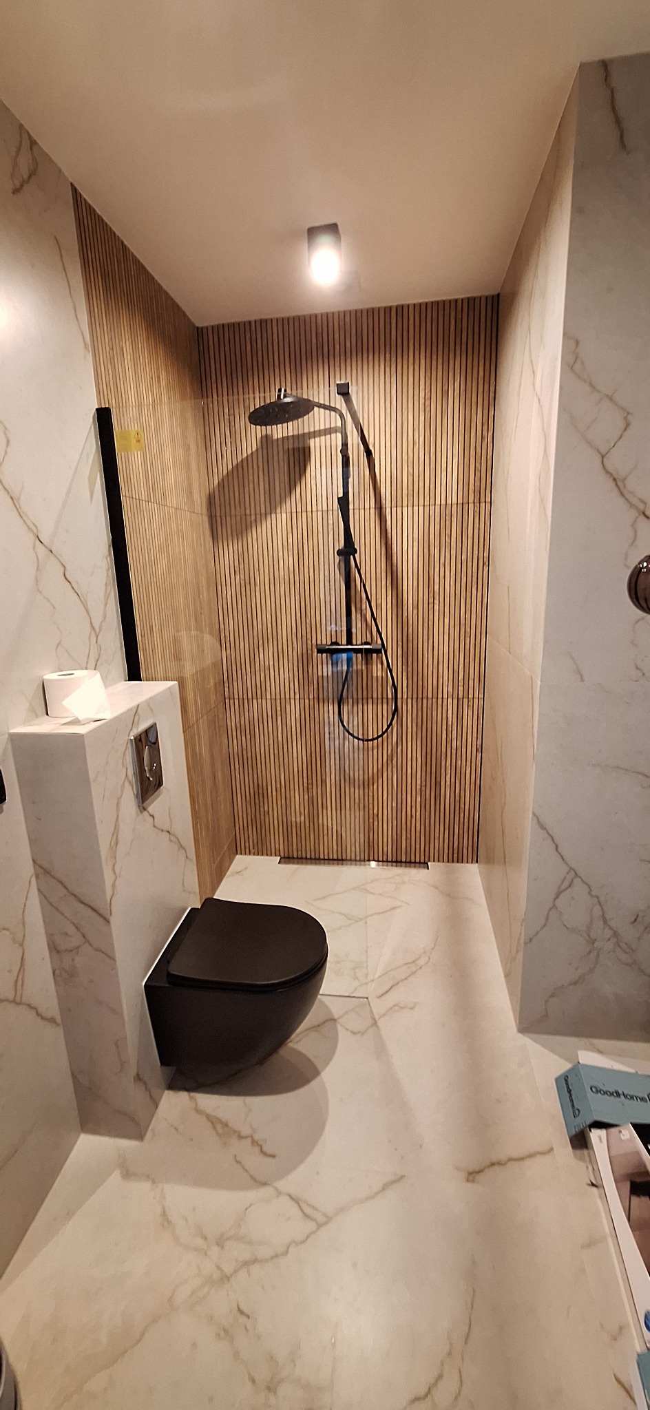 Nowoczesna łazienka z czarną miską WC i deszczownicą na tle drewnianej ściany. Marmurowe płytki podłogowe i ścienne. Minimalistyczny design.