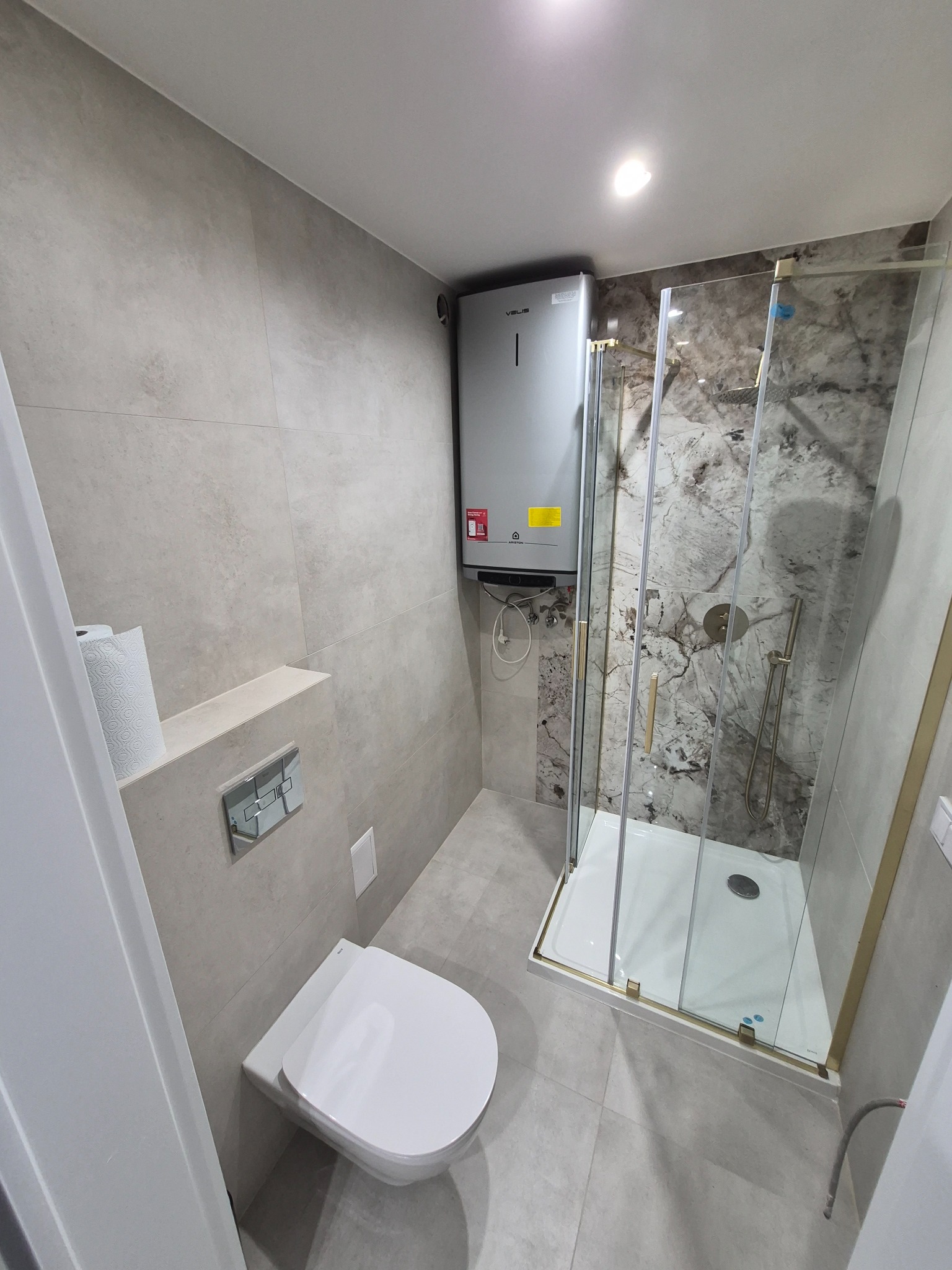 Nowoczesna łazienka z kabiną prysznicową w złotej ramie, podwieszaną toaletą i szarymi płytkami. Na ścianie zamontowany bojler. Minimalistyczny design.