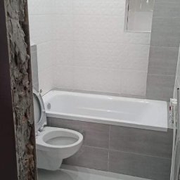 Wykończona łazienka z biało-szarą glazurą, wanną i toaletą. Widoczne detale wykończeniowe i oświetlenie sufitowe. Fragment surowej ściany po lewej stronie kadru.