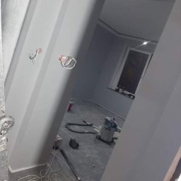 Świeżo pomalowane na szaro wnętrze pokoju z widocznymi przewodami elektrycznymi, odkurzaczem i kablem na podłodze. Widok zza framugi drzwi.