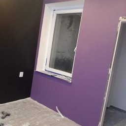 Ściana pomalowana na fioletowo z oknem i fragmentem czarnej ściany. Na podłodze widoczne narzędzia malarskie. Ukończone malowanie pokoju.