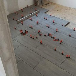 Układanie szarych płytek podłogowych z użyciem klipsów poziomujących w trakcie remontu. Widoczne narzędzia i materiały budowlane. Surowe ściany w tle.