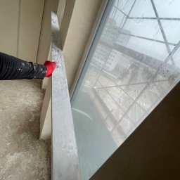 Kamil zyskowski - Ręka w czerwonej rękawicy wygładza świeżo nałożony tynk przy oknie. Widoczne rusztowania za oknem i surowy beton na podłodze. Praca w toku.
