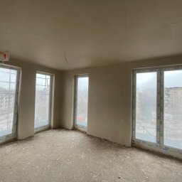 Kamil zyskowski - Wnętrze po tynkowaniu maszynowym: gładkie, jasne ściany w surowym stanie, trzy okna z widokiem na miasto. Przygotowane do dalszych prac wykończeniowych.