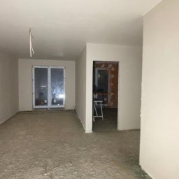 Kamil zyskowski - Wnętrze po nałożeniu tynków: gładkie, jasne ściany, surowa podłoga, okno z widokiem na nocne miasto, widoczne kable i przygotowane otwory drzwiowe.
