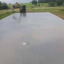 Arkadiusz Nosidlak ResinMix - Świeżo wylana, gładka posadzka z widocznymi dylatacjami i kratkami odpływowymi. W tle koparka i zielony krajobraz. Etap prac budowlanych.