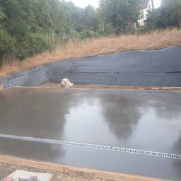 Arkadiusz Nosidlak ResinMix - Świeżo wylana, gładka posadzka przemysłowa z odpływem liniowym, otoczona drewnianym szalunkiem. W tle skarpa zabezpieczona czarną folią i zieleń.