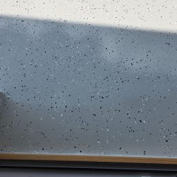 Arkadiusz Nosidlak ResinMix - Jasnoszara posadzka z drobnymi, czarnymi i białymi elementami dekoracyjnymi, wykończona drewnianą listwą przypodłogową i fragmentem ściany w tle.