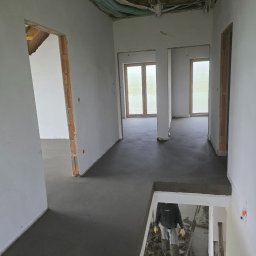 Arkadiusz Nosidlak ResinMix - Wnętrze w trakcie wylewania posadzki: świeży, szary beton na podłodze, widoczne instalacje pod sufitem, pracownik w kaloszach wchodzi po schodach. Surowy stan wykończenia.
