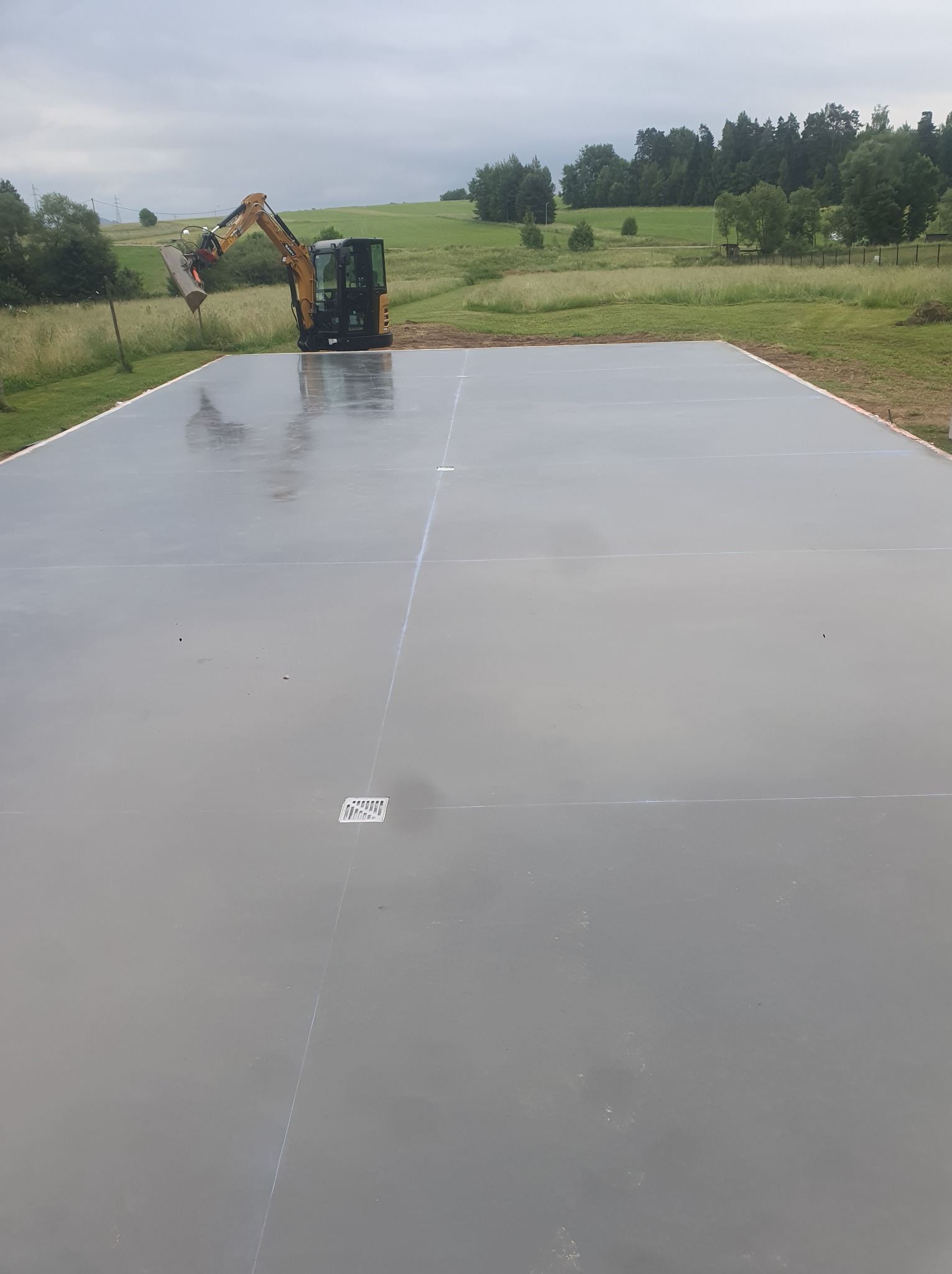 Świeżo wylana, gładka posadzka z widocznymi dylatacjami i kratkami odpływowymi. W tle koparka i zielony krajobraz. Etap prac budowlanych.