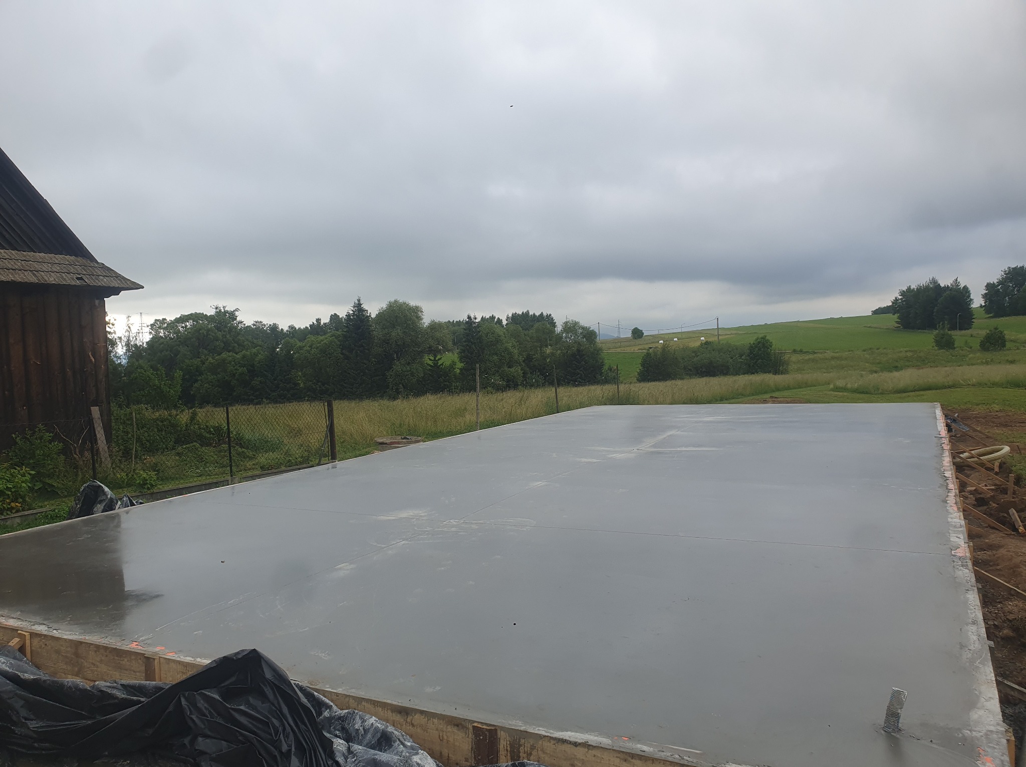 Świeżo wylana, gładka posadzka na zewnątrz, otoczona drewnianym szalunkiem. W tle zielony krajobraz i pochmurne niebo. Widok z góry, lekko z ukosa.