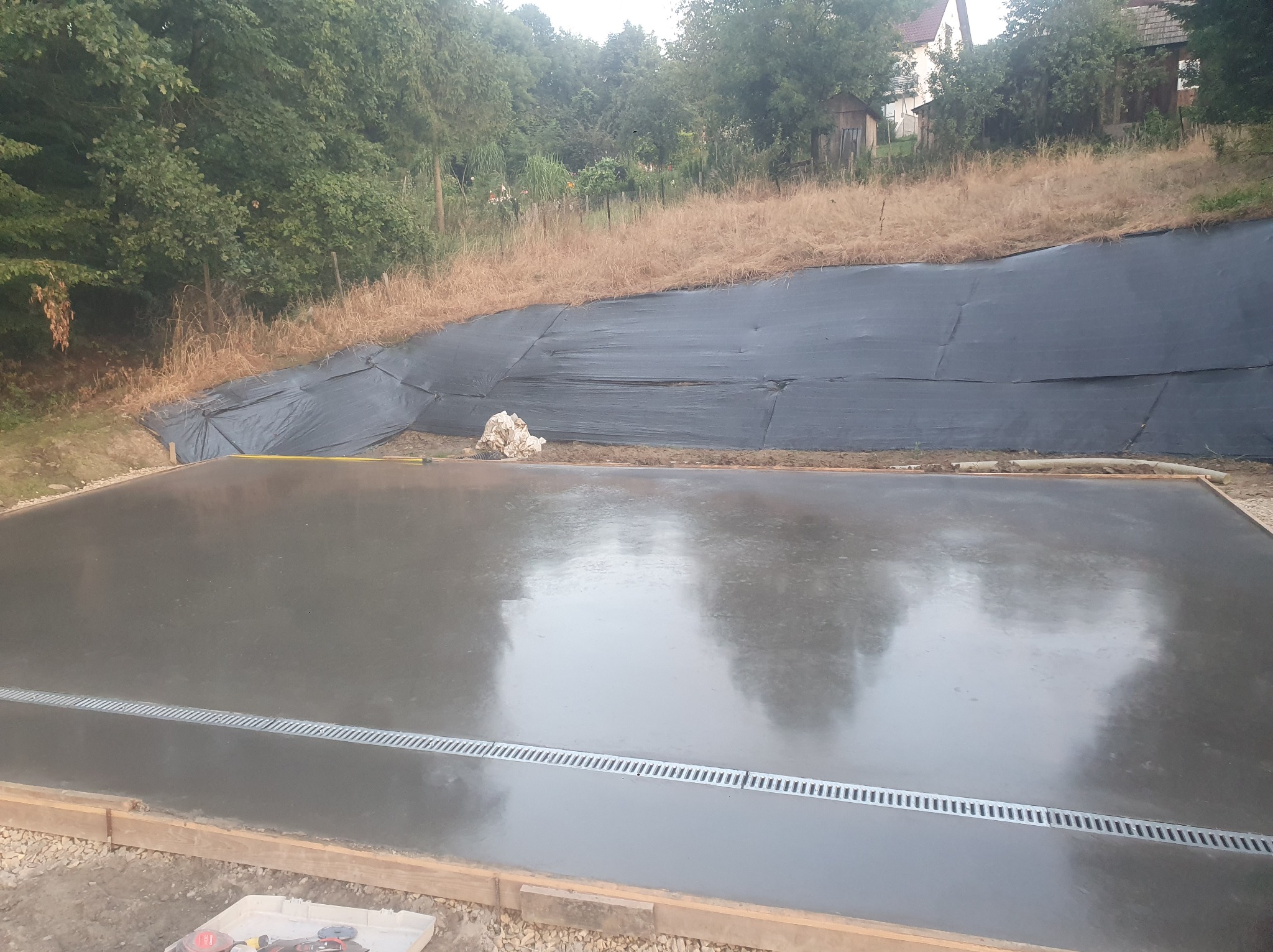 Świeżo wylana, gładka posadzka przemysłowa z odpływem liniowym, otoczona drewnianym szalunkiem. W tle skarpa zabezpieczona czarną folią i zieleń.