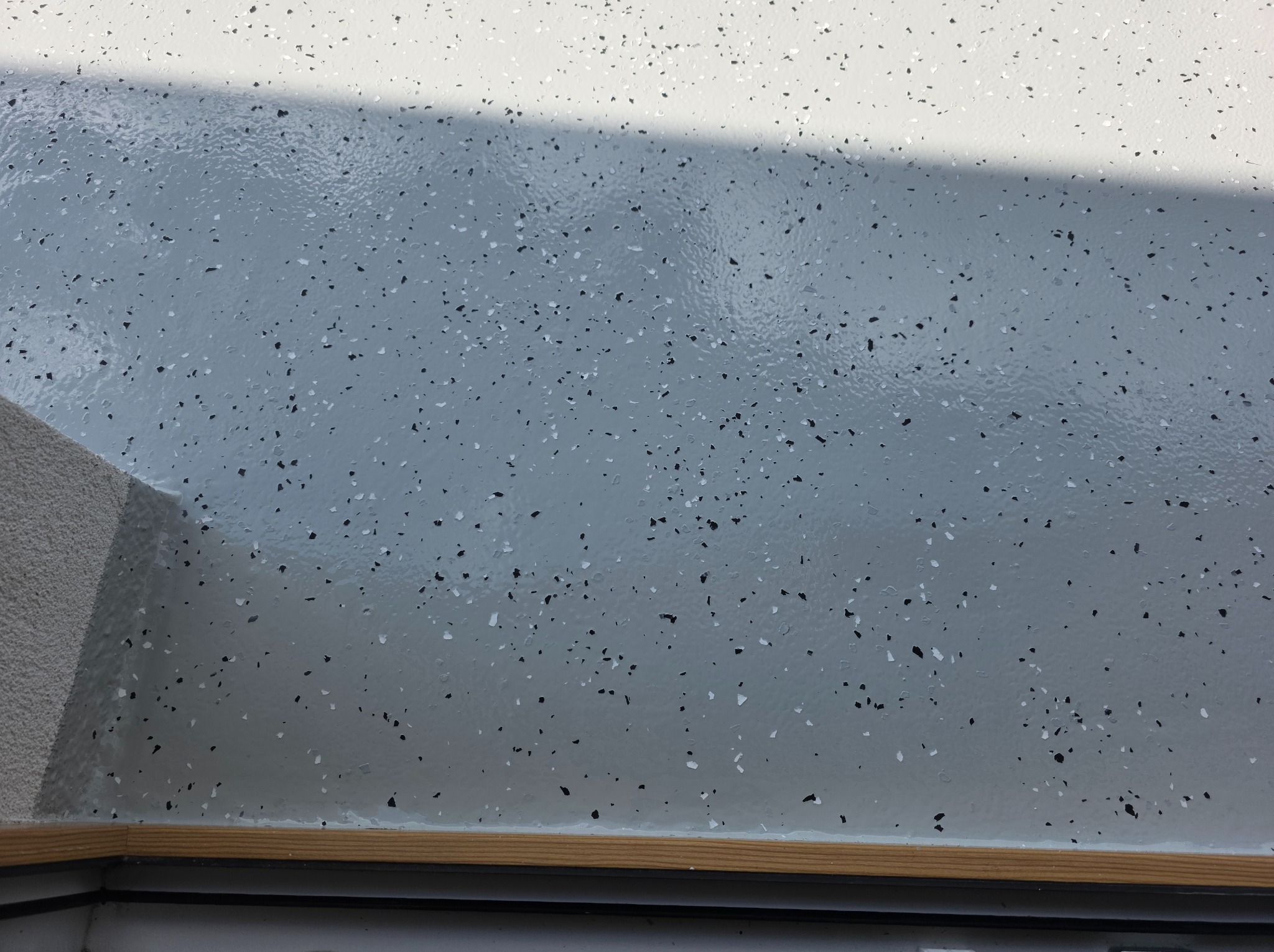 Jasnoszara posadzka z drobnymi, czarnymi i białymi elementami dekoracyjnymi, wykończona drewnianą listwą przypodłogową i fragmentem ściany w tle.