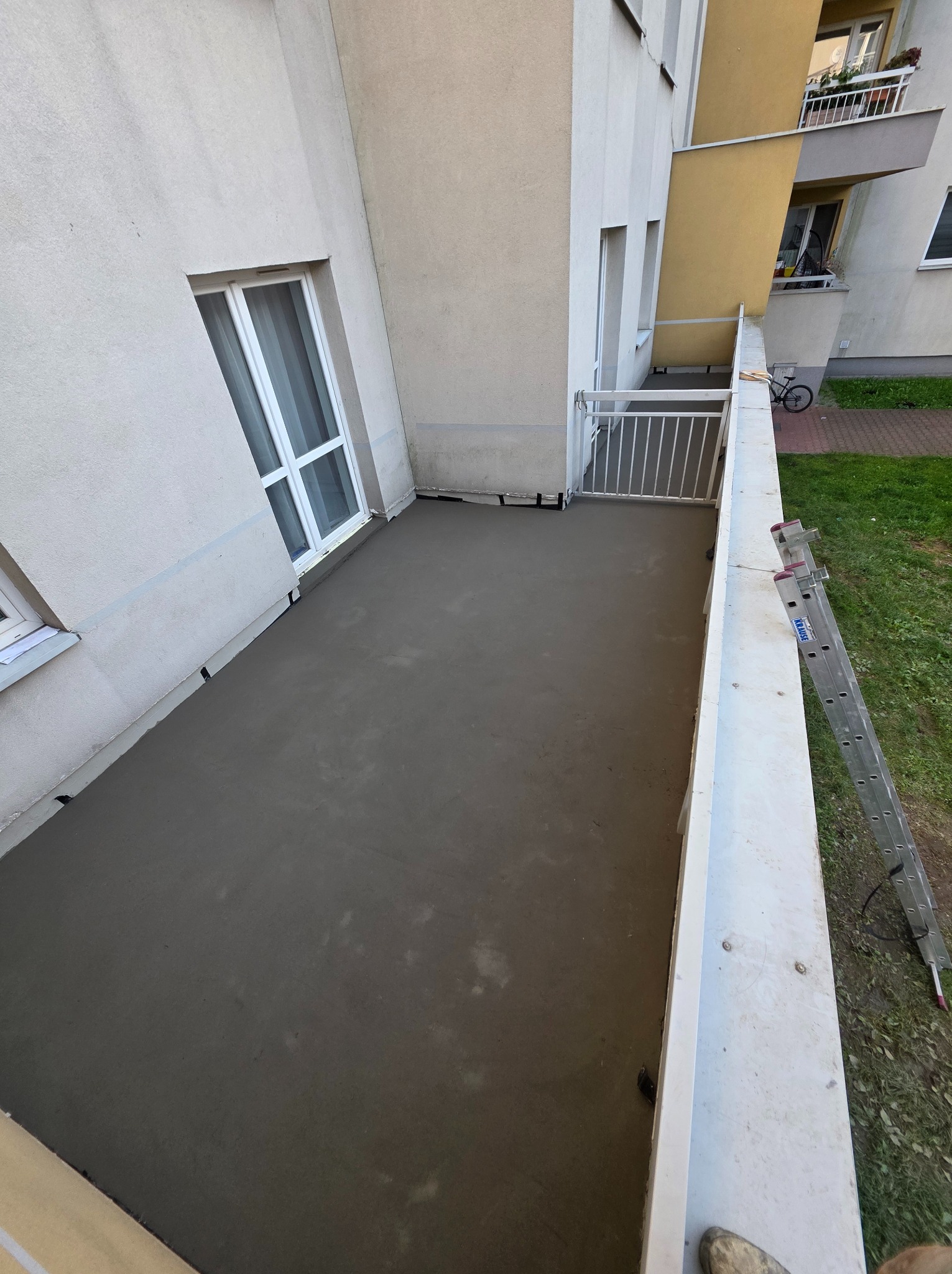 Wylana, gładka posadzka na balkonie w bloku. Widoczna drabina oparta o barierkę. Szara, jednolita powierzchnia. Nowa wylewka na balkonie.