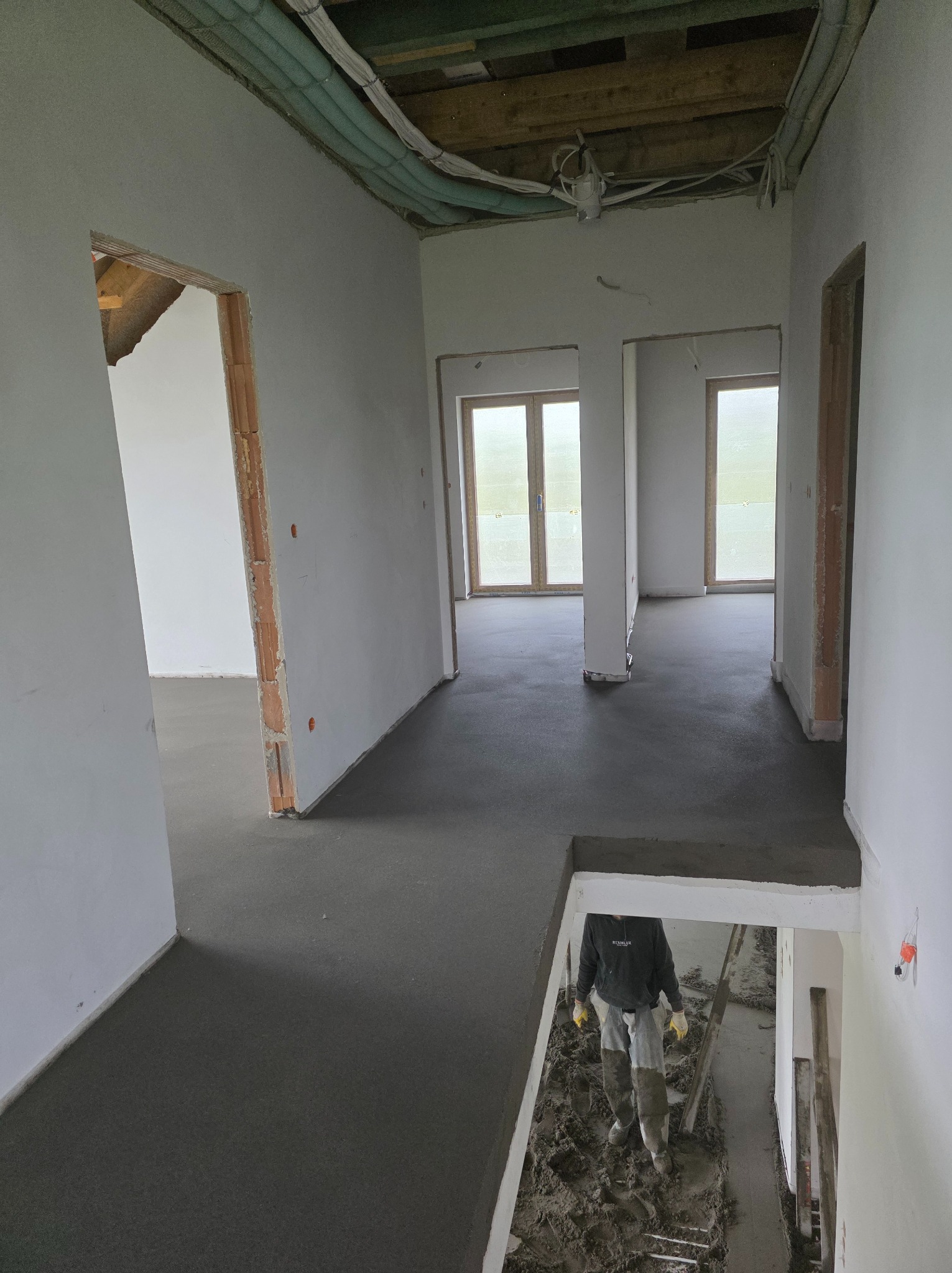 Wnętrze w trakcie wylewania posadzki: świeży, szary beton na podłodze, widoczne instalacje pod sufitem, pracownik w kaloszach wchodzi po schodach. Surowy stan wykończenia.