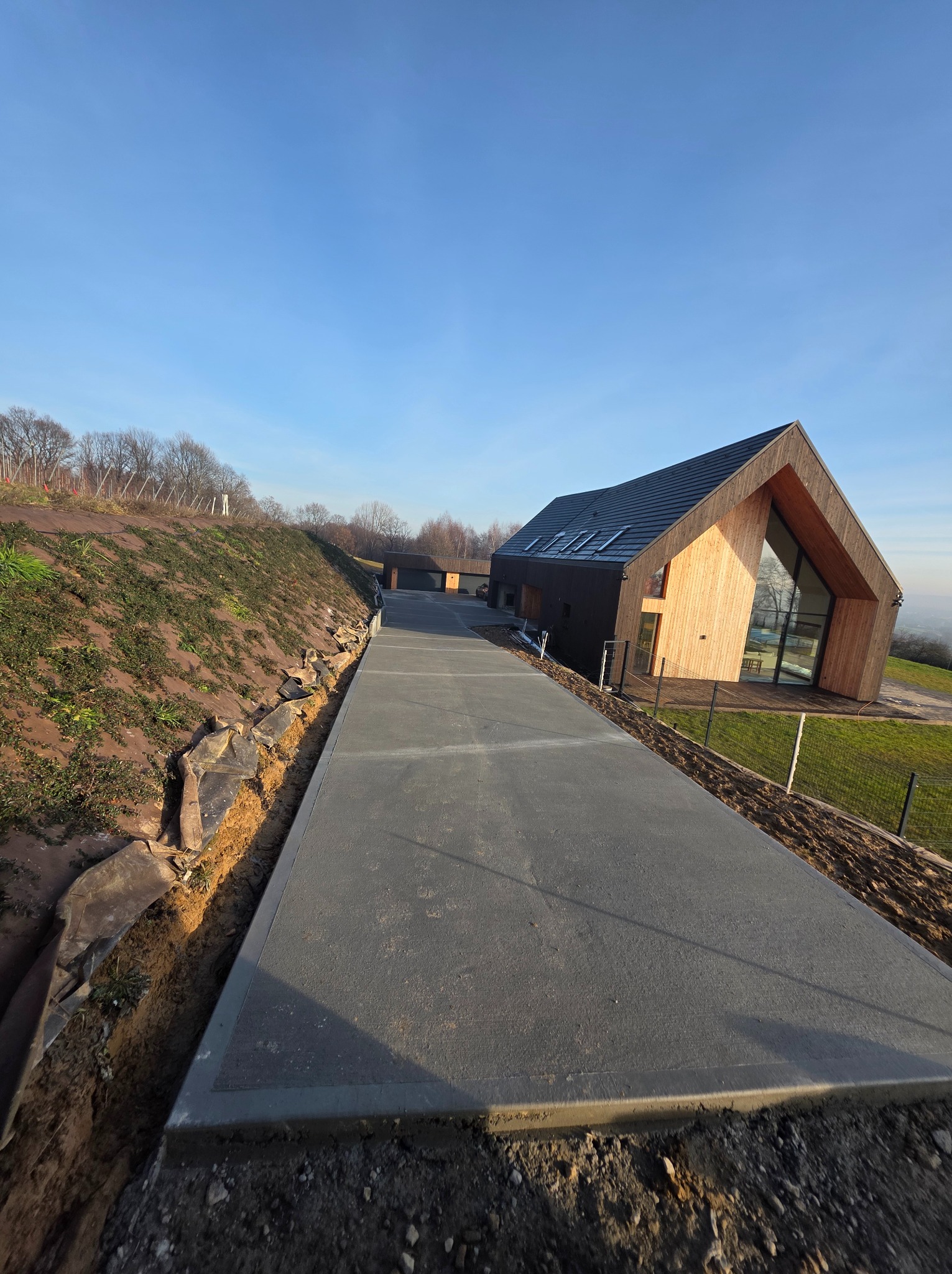 Długa, szara droga dojazdowa z betonu prowadząca do nowoczesnego domu z drewnianą fasadą na wzgórzu, widok z dołu, błękitne niebo w tle.