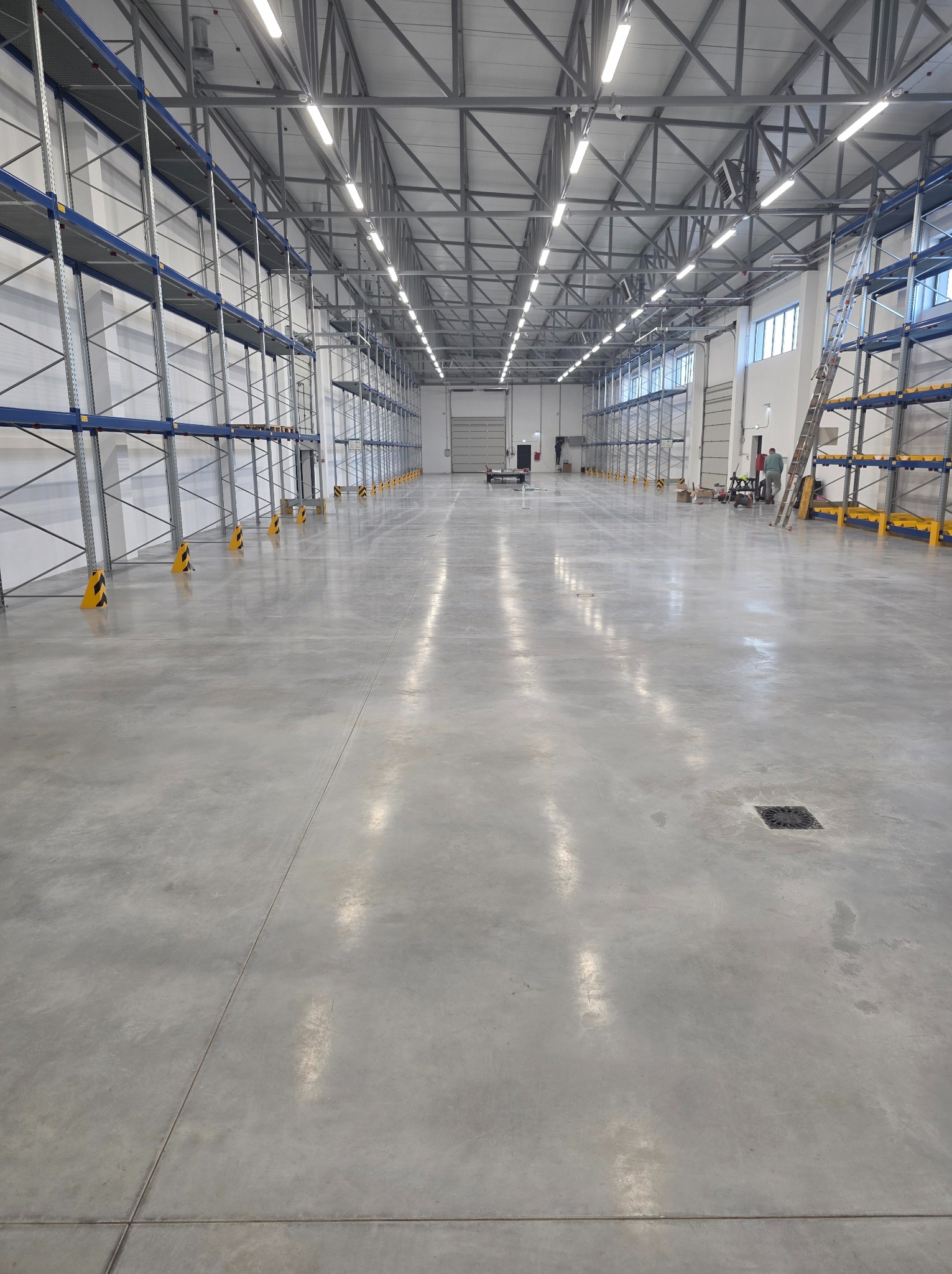 Wnętrze hali magazynowej z szarą, gładką posadzką przemysłową, regałami wzdłuż ścian i oświetleniem sufitowym. Widoczny właz rewizyjny w podłodze i drabina oparta o regał.