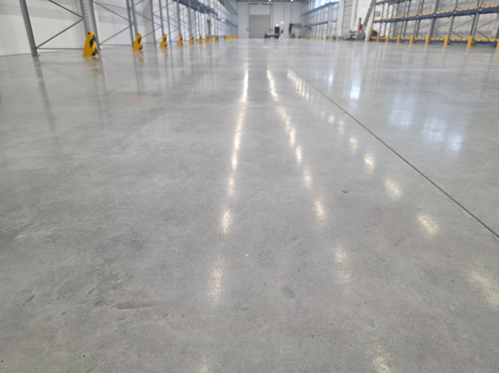 Błyszcząca, szara posadzka przemysłowa w hali magazynowej z żółto-czarnymi słupami zabezpieczającymi i regałami w tle. Widok z niskiej perspektywy.