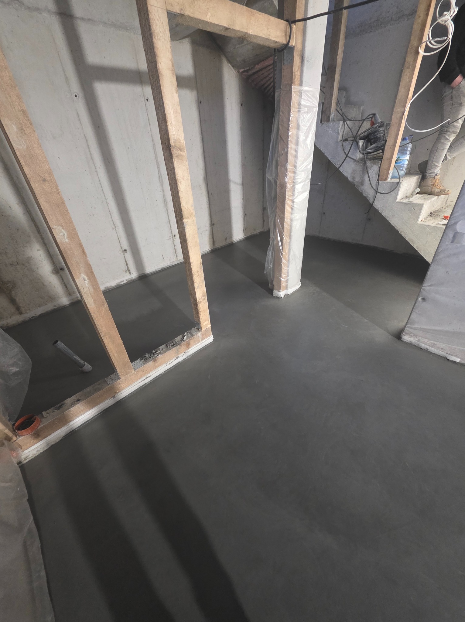 Wylana, gładka, szara posadzka w surowym wnętrzu z betonowymi ścianami i drewnianą konstrukcją. Widoczne schody w tle i częściowo zakryte folią elementy.