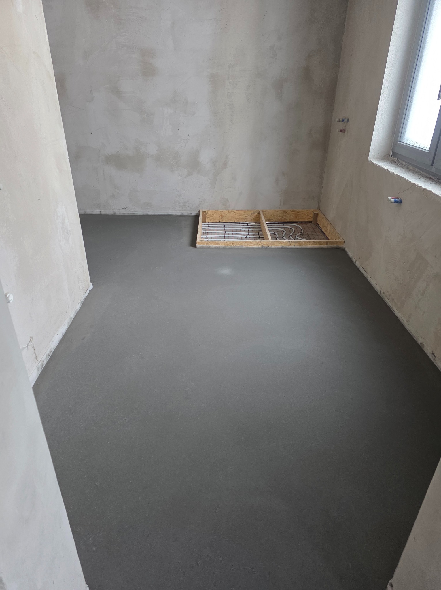 Wylana, szara posadzka w surowym wnętrzu z widocznym fragmentem instalacji ogrzewania podłogowego w drewnianej ramie. Ściany w stanie deweloperskim, okno z prawej strony.