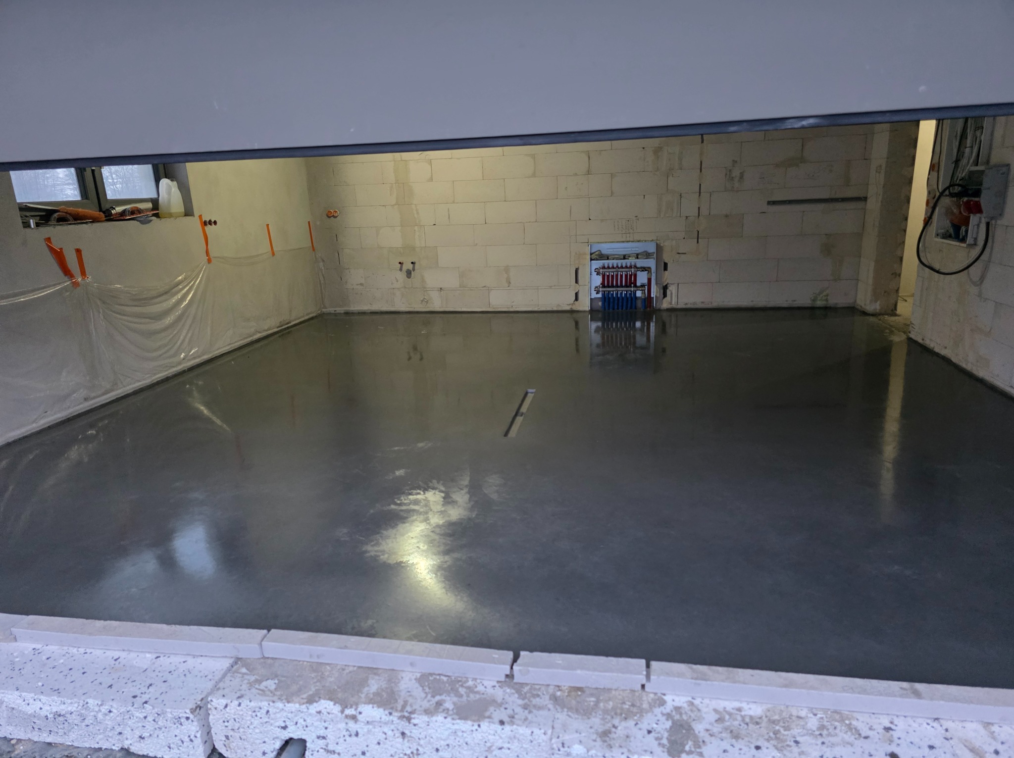 Świeżo wylana, gładka, szara posadzka w surowym wnętrzu z białych bloczków. Widoczna instalacja grzewcza na ścianie i folia ochronna. Częściowo opuszczona brama garażowa.