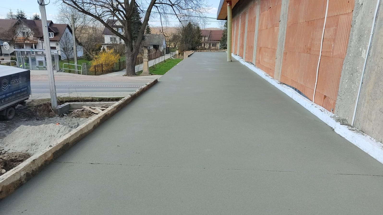 Świeżo wylana, gładka, szara posadzka na zewnątrz budynku w trakcie realizacji. Widoczna ściana z cegły, krawężnik i fragment ulicy z budynkiem w tle.