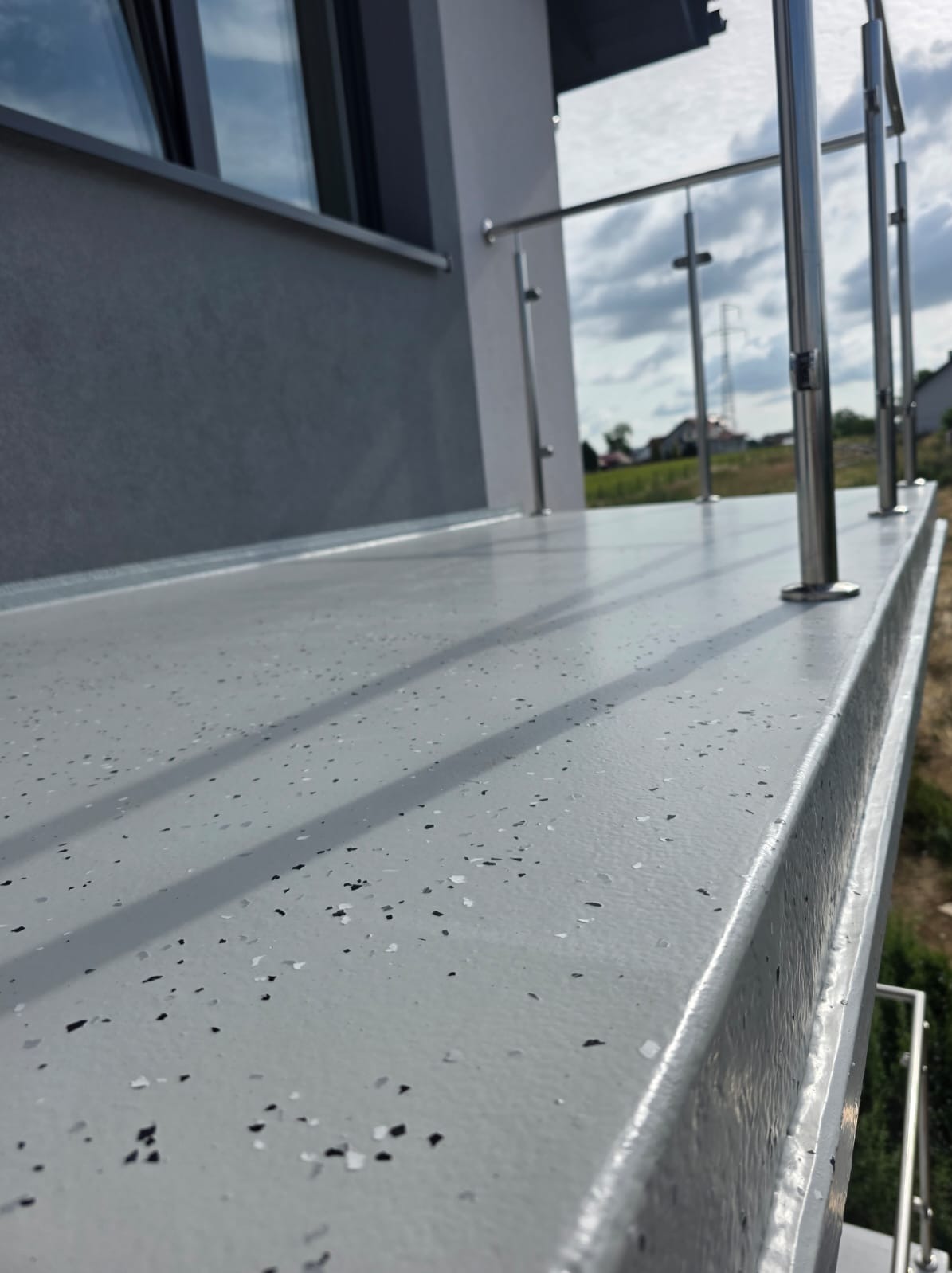 Szara, gładka powierzchnia balkonu z żywicy epoksydowej, z widocznymi drobnymi, czarnymi i białymi elementami. Metalowe barierki w tle, budynek i zieleń w oddali.