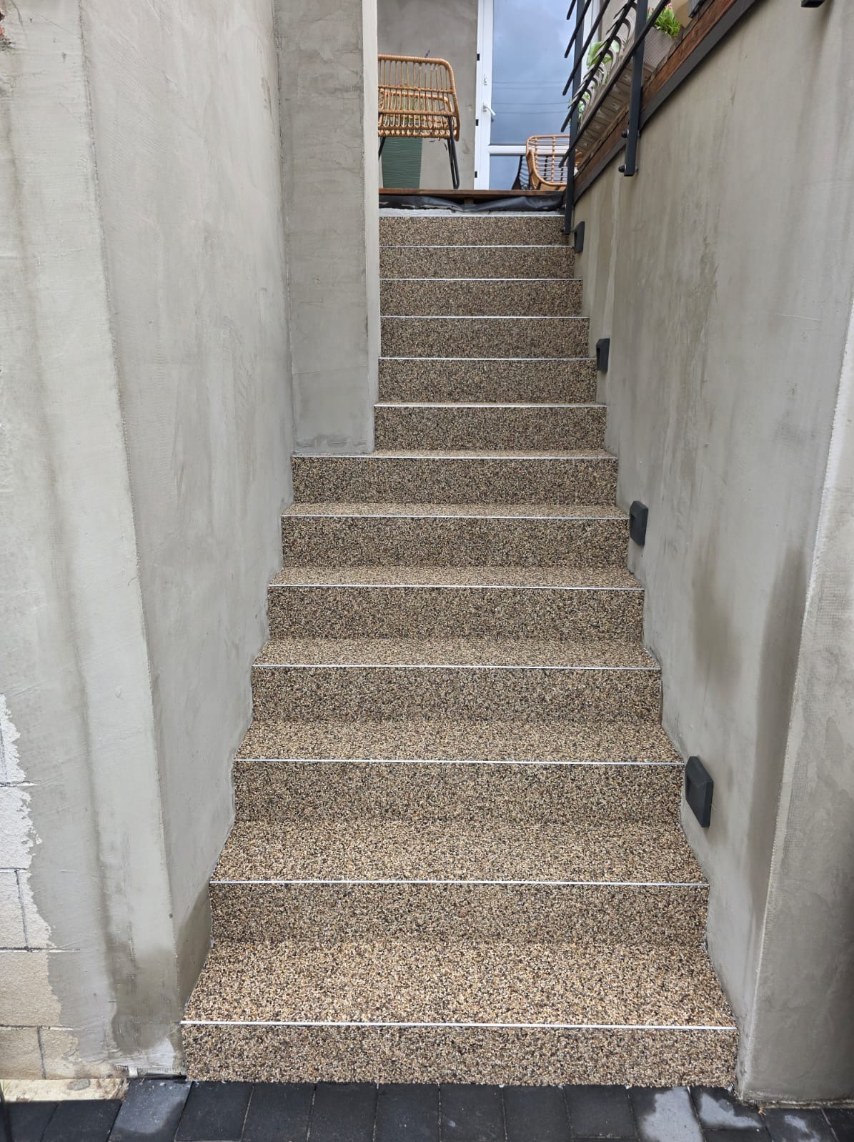 Schody zewnętrzne z żywicy epoksydowej w odcieniach beżu i brązu, wykończone białymi listwami, prowadzące na taras z rattanowymi krzesłami. Surowe, betonowe ściany.