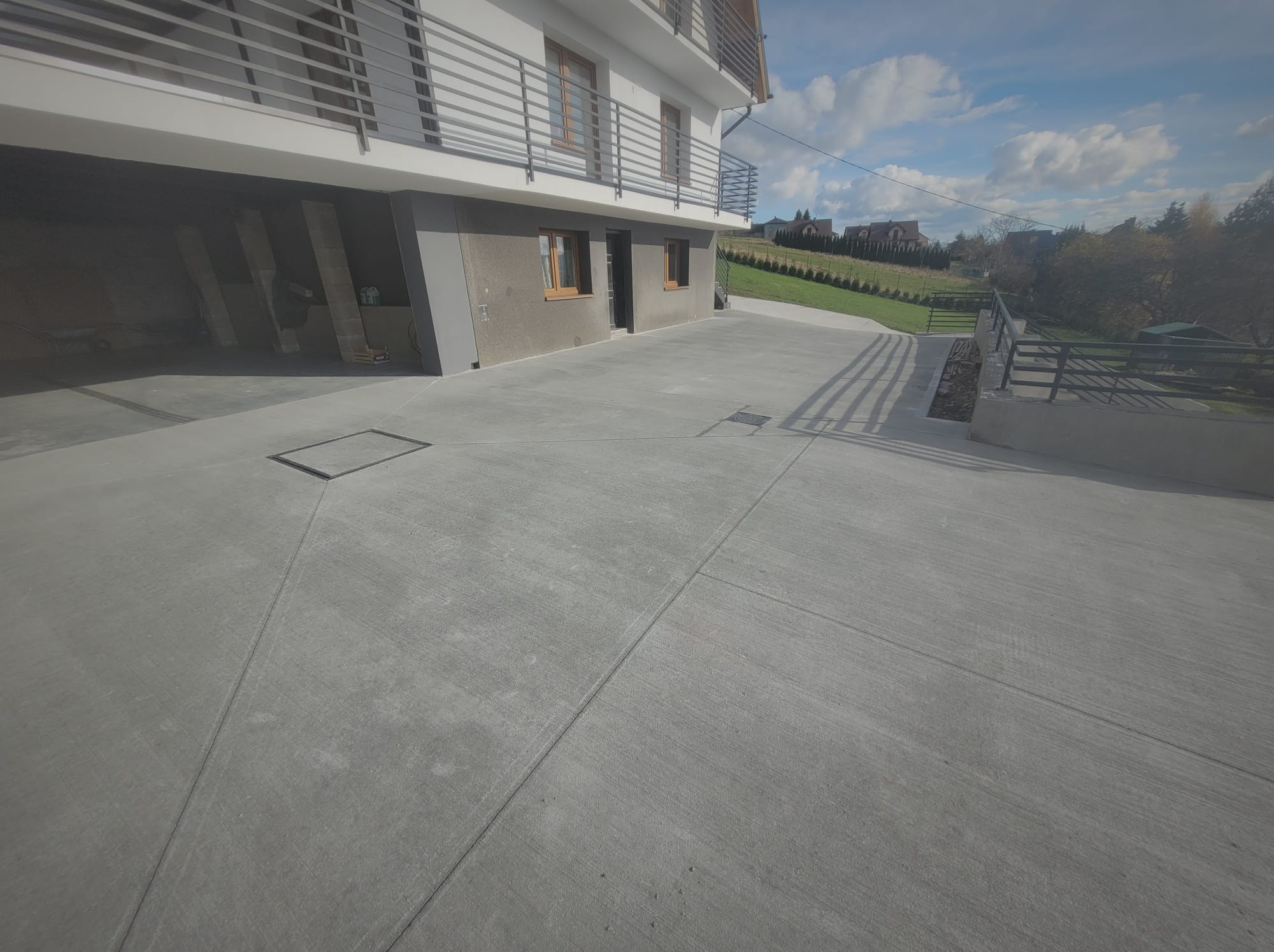 Szara, gładka posadzka przemysłowa na zewnątrz budynku z widocznymi liniami dylatacyjnymi i kratką odpływową. Tło stanowi dom z balkonem i zielony teren.
