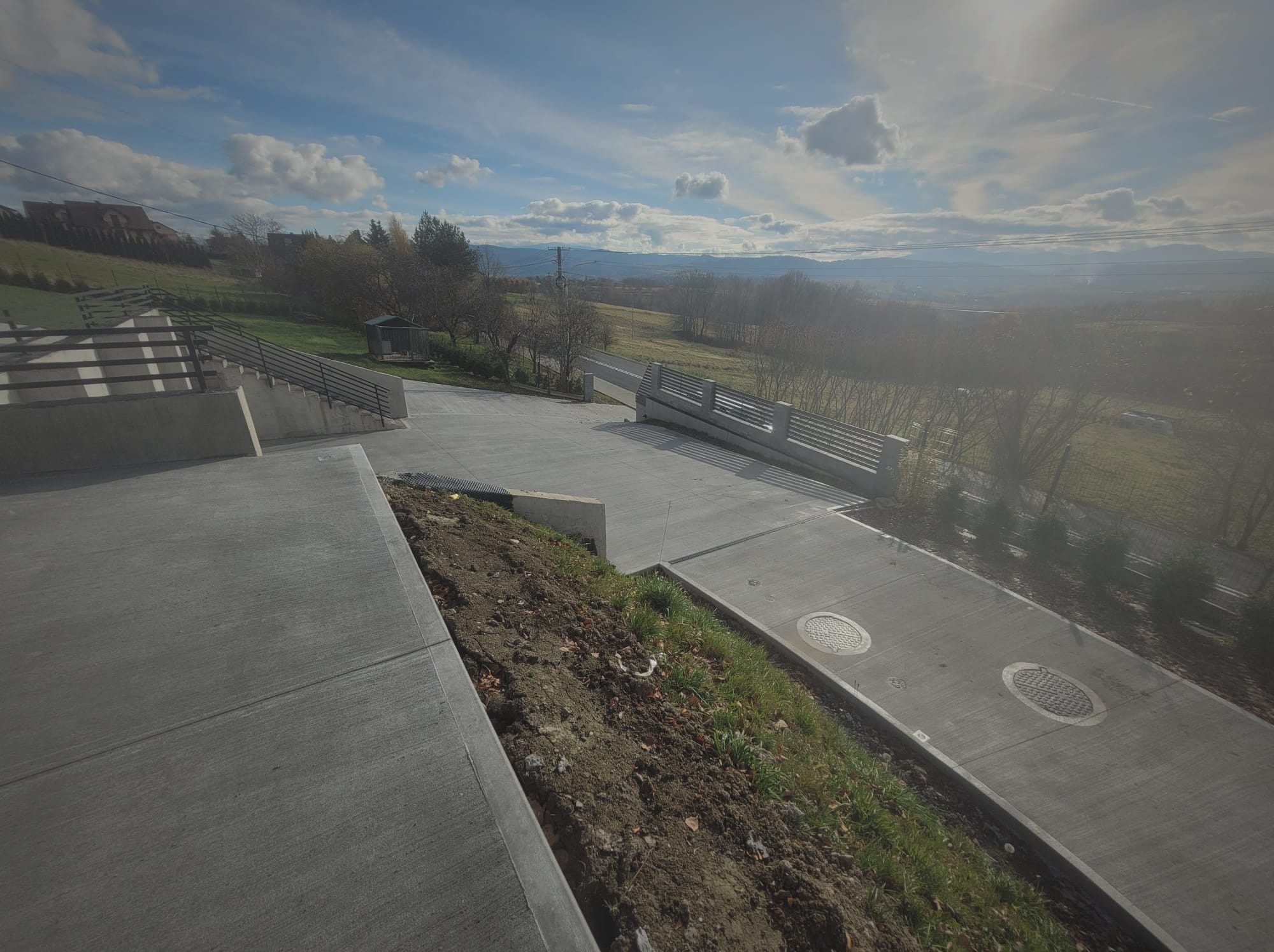 Betonowa droga dojazdowa z włazami kanalizacyjnymi, otoczona zielenią i widokiem na górzysty krajobraz w tle, w słoneczny dzień.