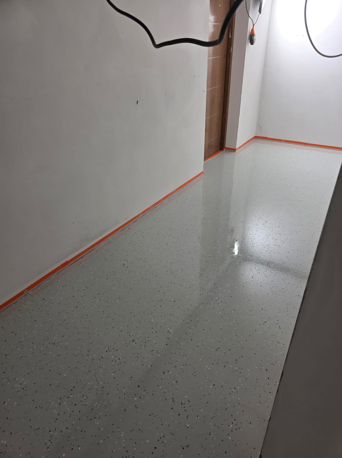 Błyszcząca, szara posadzka żywiczna z drobnymi, ciemnymi inkluzjami, zabezpieczona pomarańczową taśmą malarską przy ścianach w jasnym pomieszczeniu.