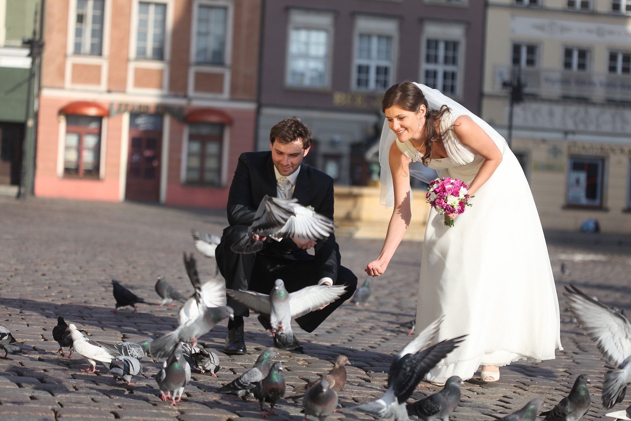 Uśmiechnięta para młoda w strojach ślubnych karmiąca gołębie na brukowanym placu miejskim w słoneczny dzień. W tle budynki z oknami.