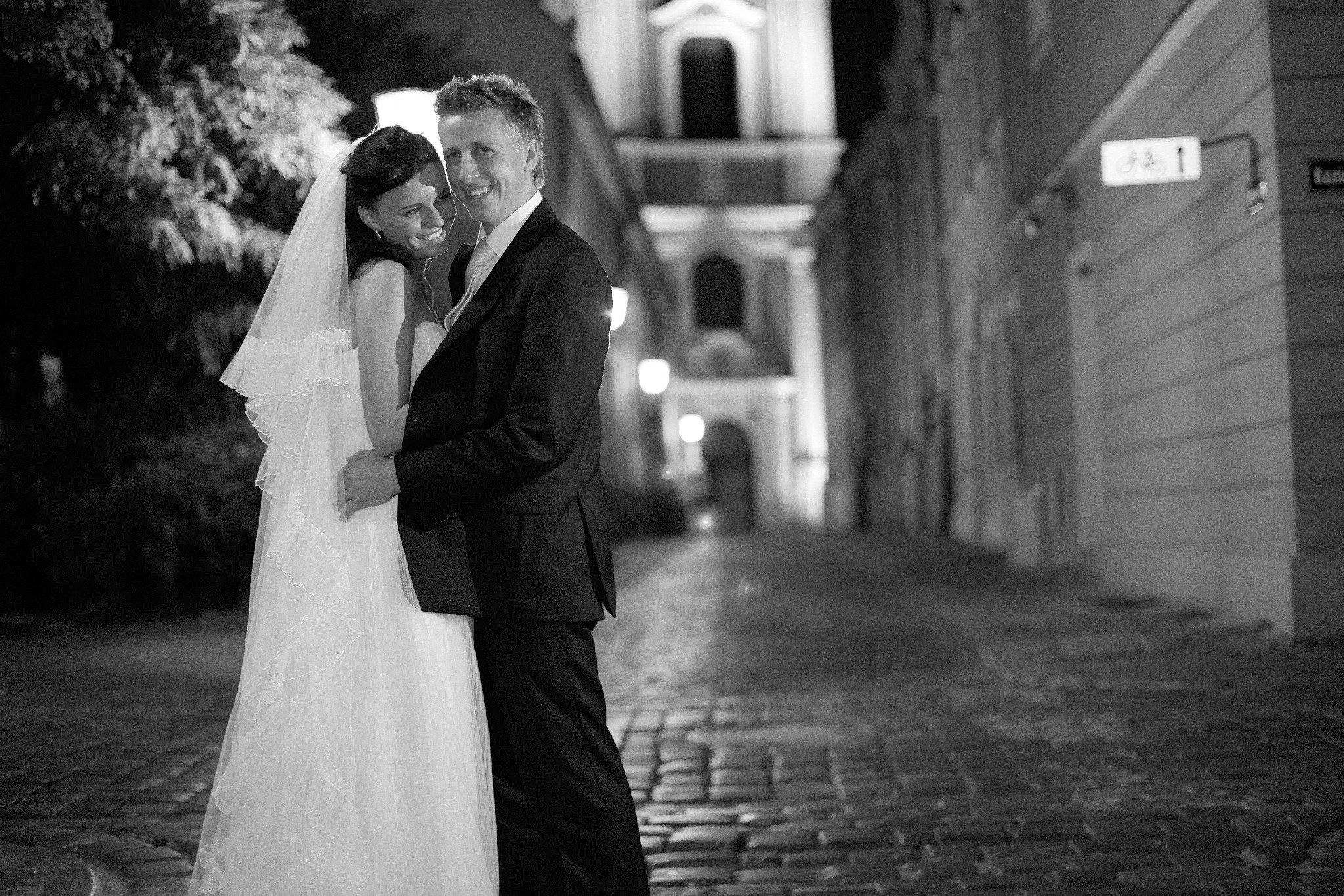Czarno-białe zdjęcie uśmiechniętej pary młodej w objęciach na brukowanej uliczce, nocą, z budynkami w tle. Panna młoda w welonie, pan młody w garniturze.