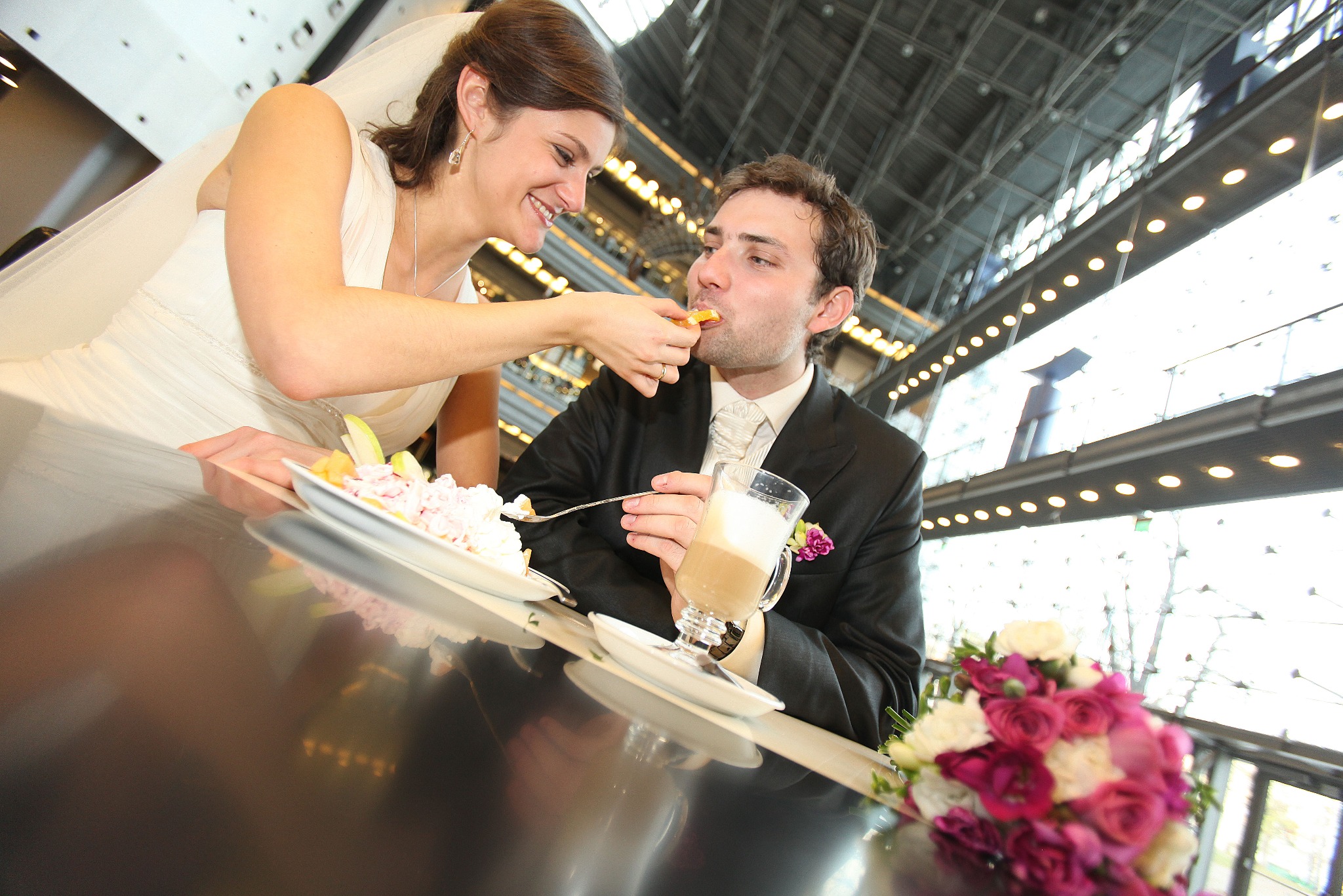 Panna młoda karmi pana młodego ciastem przy stole z bukietem. Para w strojach ślubnych w jasnym wnętrzu z widoczną konstrukcją sufitu.