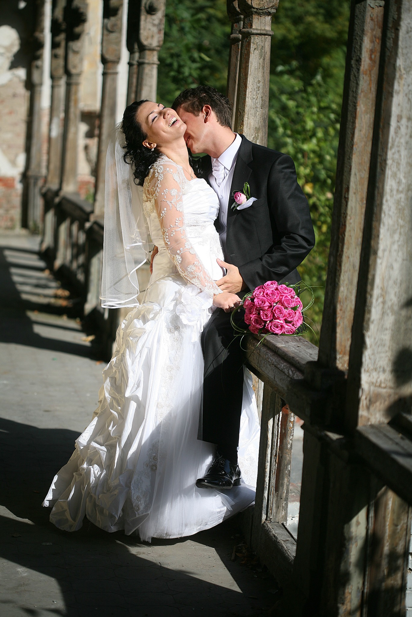 Uśmiechnięta panna młoda w koronkowej sukni i welonie, obejmowana przez pana młodego w czarnym garniturze, z bukietem różowych róż na tle kolumnady.