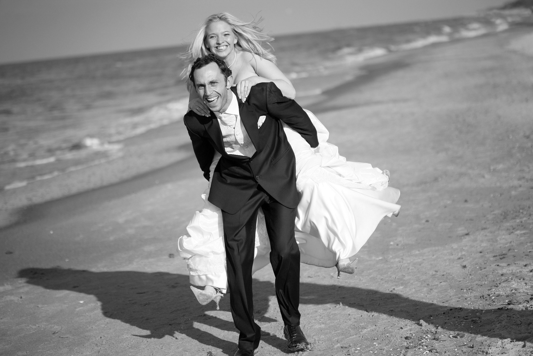 Czarno-białe zdjęcie: uśmiechnięty pan młody niesie na barana roześmianą pannę młodą na plaży, w tle morze. Dynamiczne ujęcie, wiatr we włosach.