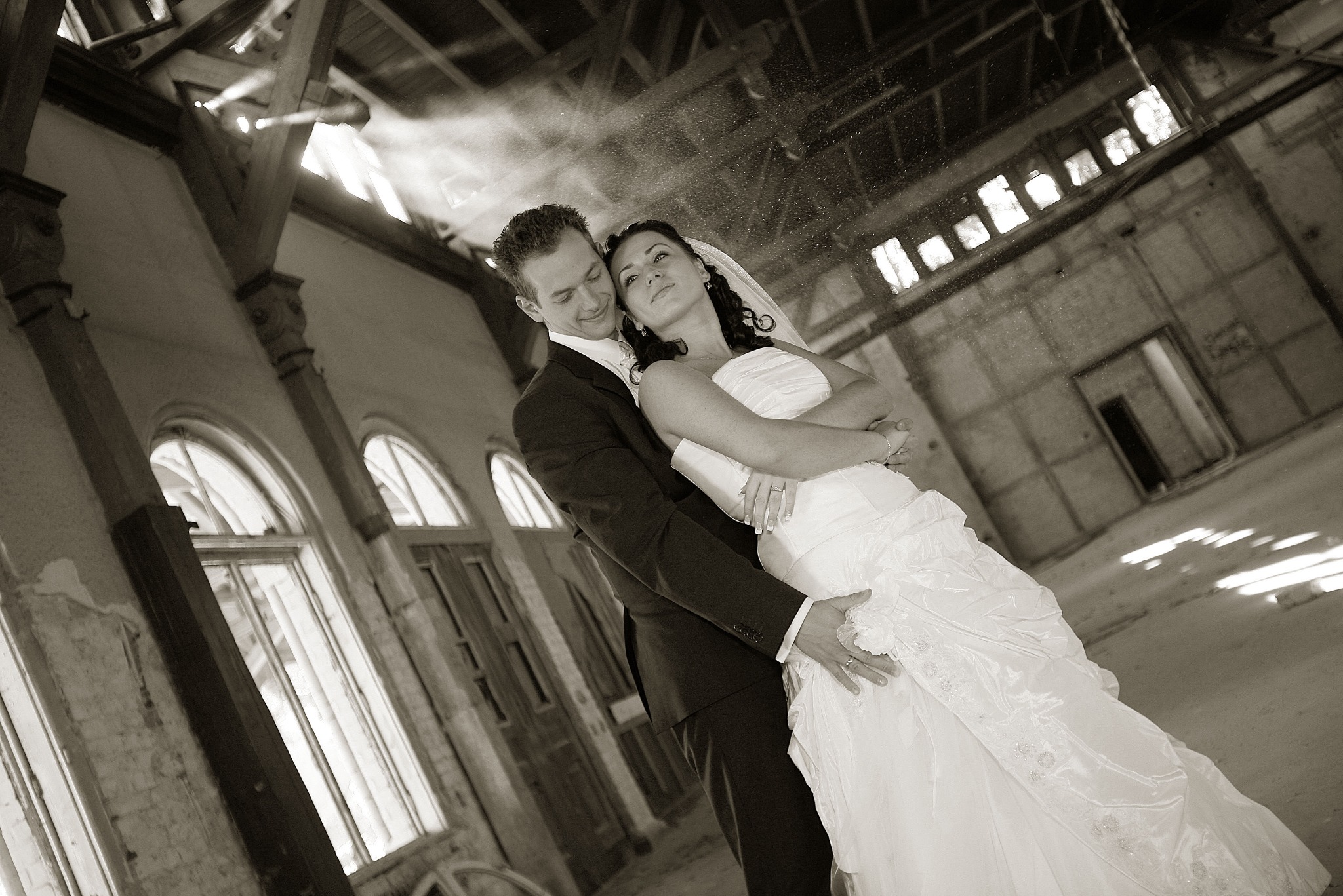 Para młoda w objęciach w opuszczonej hali z oknami łukowymi, w sepii. Romantyczna sesja ślubna w Poznaniu z efektem padającego światła.