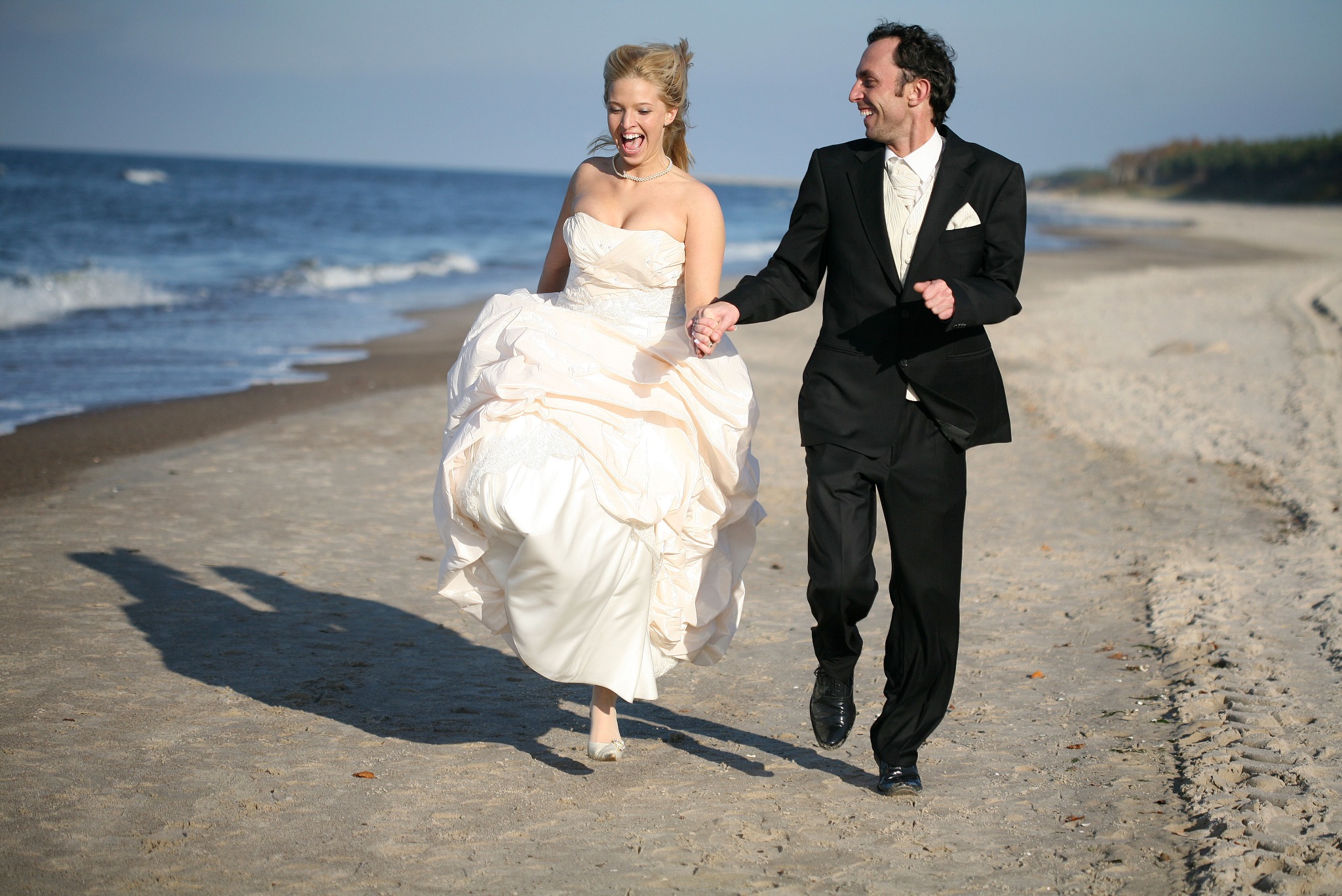 Uśmiechnięta para młoda biegnie po piaszczystej plaży, trzymając się za ręce. Panna młoda w sukni bez ramiączek, pan młody w czarnym garniturze. W tle morze i las.