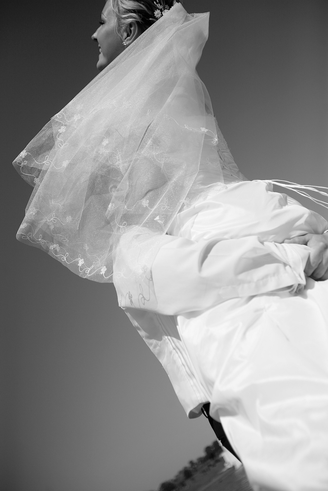 Czarno-białe zdjęcie panny młodej w sukni i welonie, uchwycone z dołu pod kątem, z widocznym niebem w tle. Delikatne zdobienia na welonie.