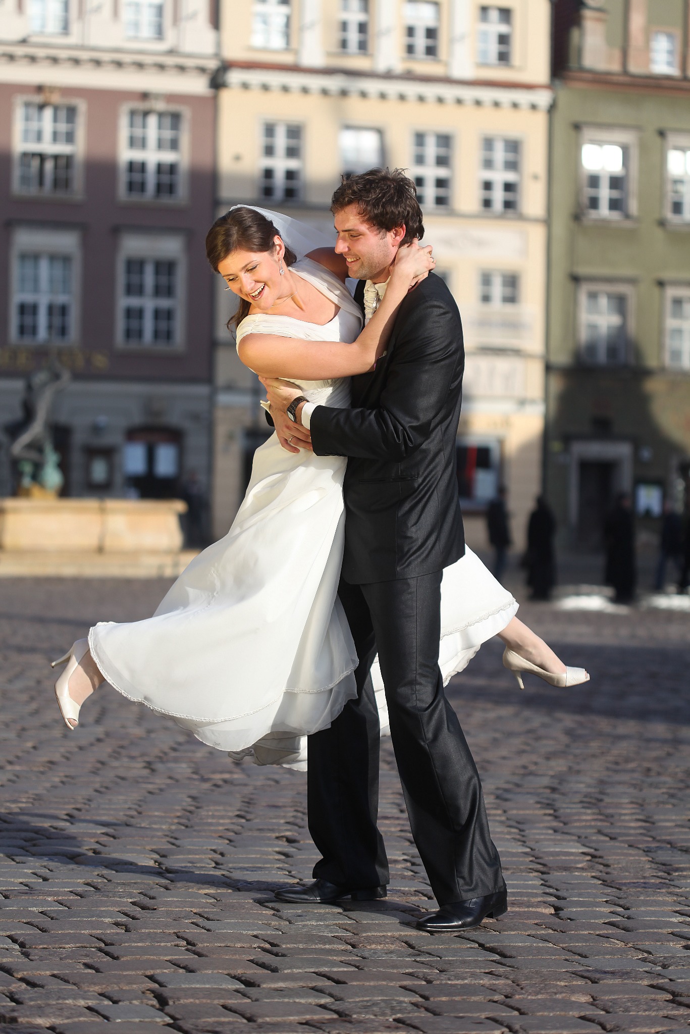 Uśmiechnięta panna młoda w białej sukni uniesiona przez pana młodego na tle kolorowych kamienic i brukowanej nawierzchni placu miejskiego w słoneczny dzień.