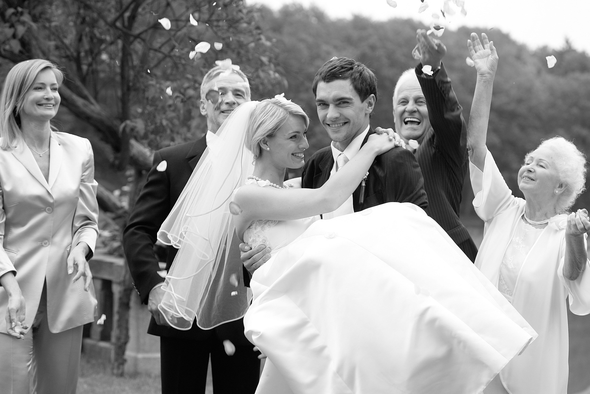 Czarno-białe zdjęcie: Uśmiechnięty pan młody trzyma na rękach pannę młodą w białej sukni, w tle goście weselni rzucający płatki kwiatów, radosna atmosfera.