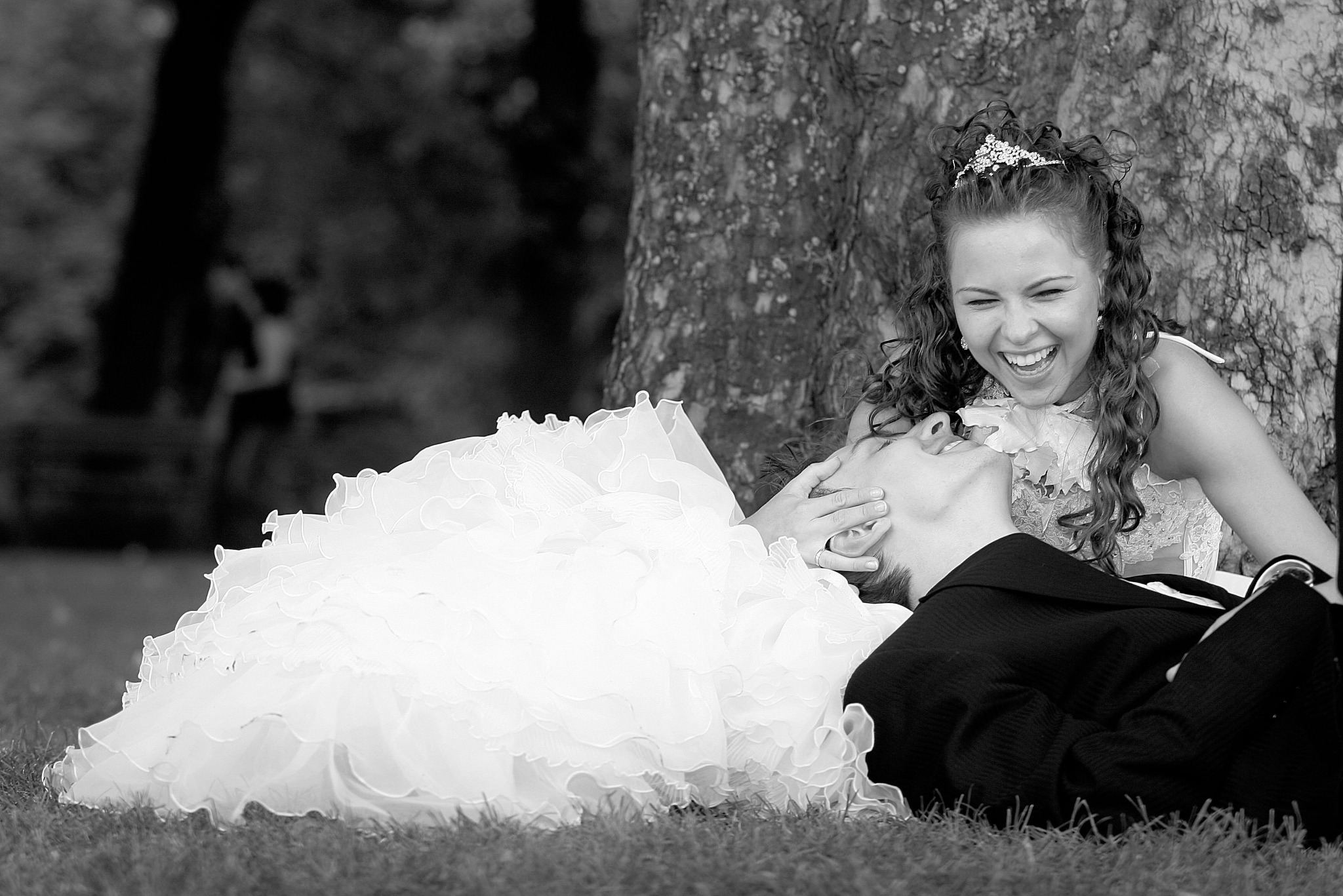 Czarno-białe zdjęcie uśmiechniętej panny młodej w tiarze, leżącej na trawie obok drzewa, z panem młodym w garniturze, który również leży na trawie i patrzy na nią.
